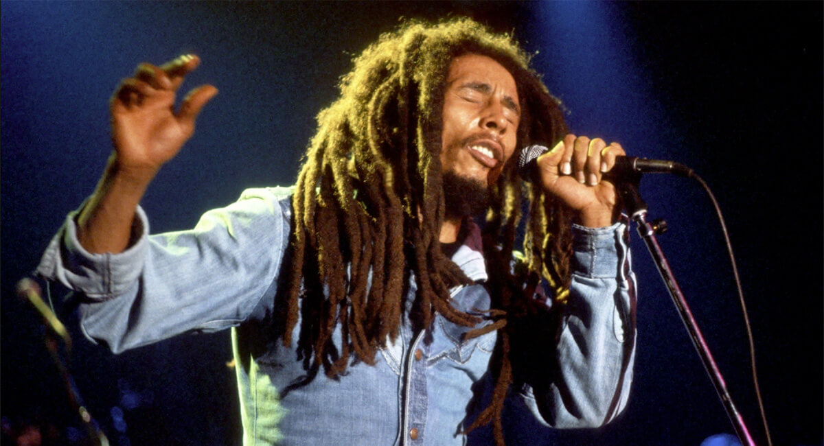 Bob Marley with thick long dread locs past his chest singing deeply into a microphone he is holdingfeatured in the Best Hair in Rock and Roll.