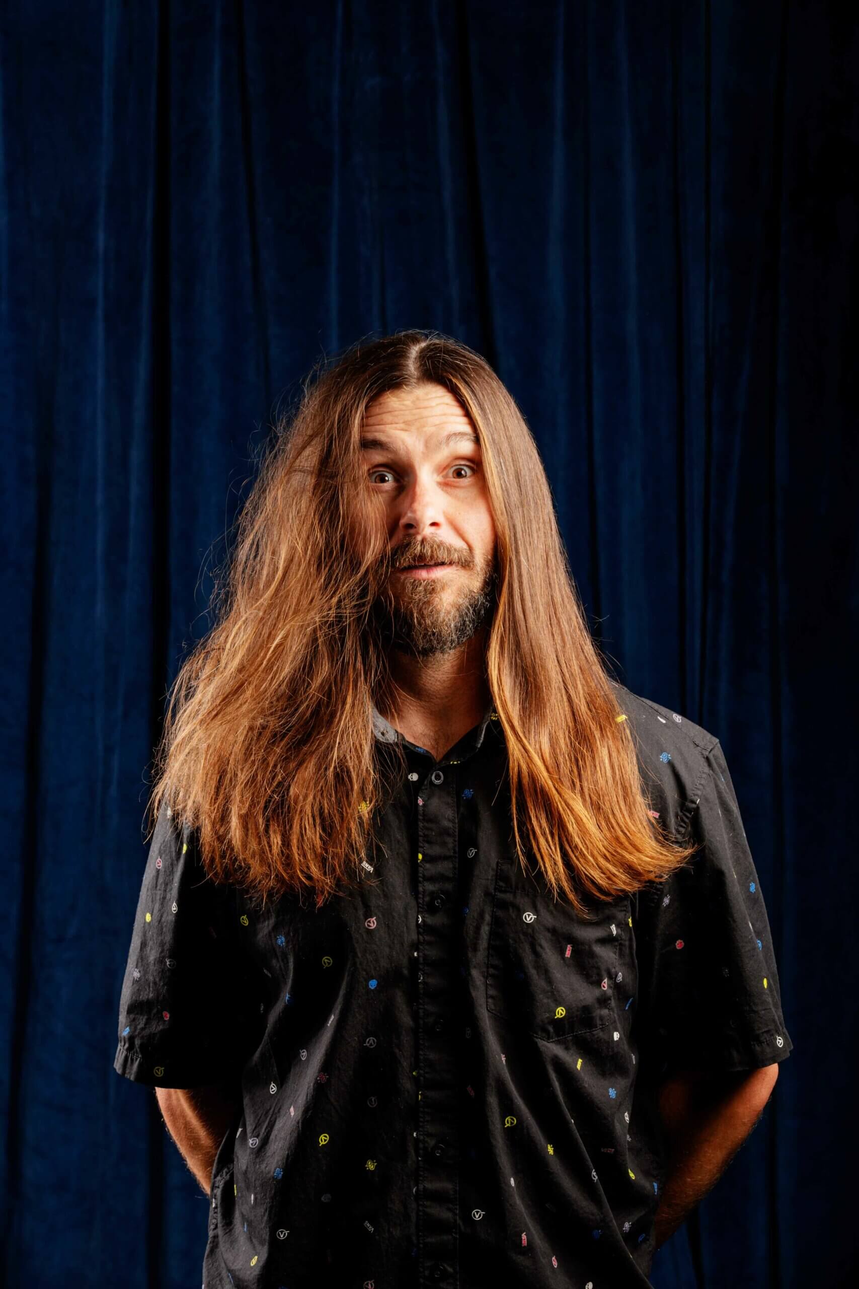 El Moreno standing in The Longhairs set. Half of his hair is perfect the other half is an absolute mess, featured in The Longhairs 2022 Status Update