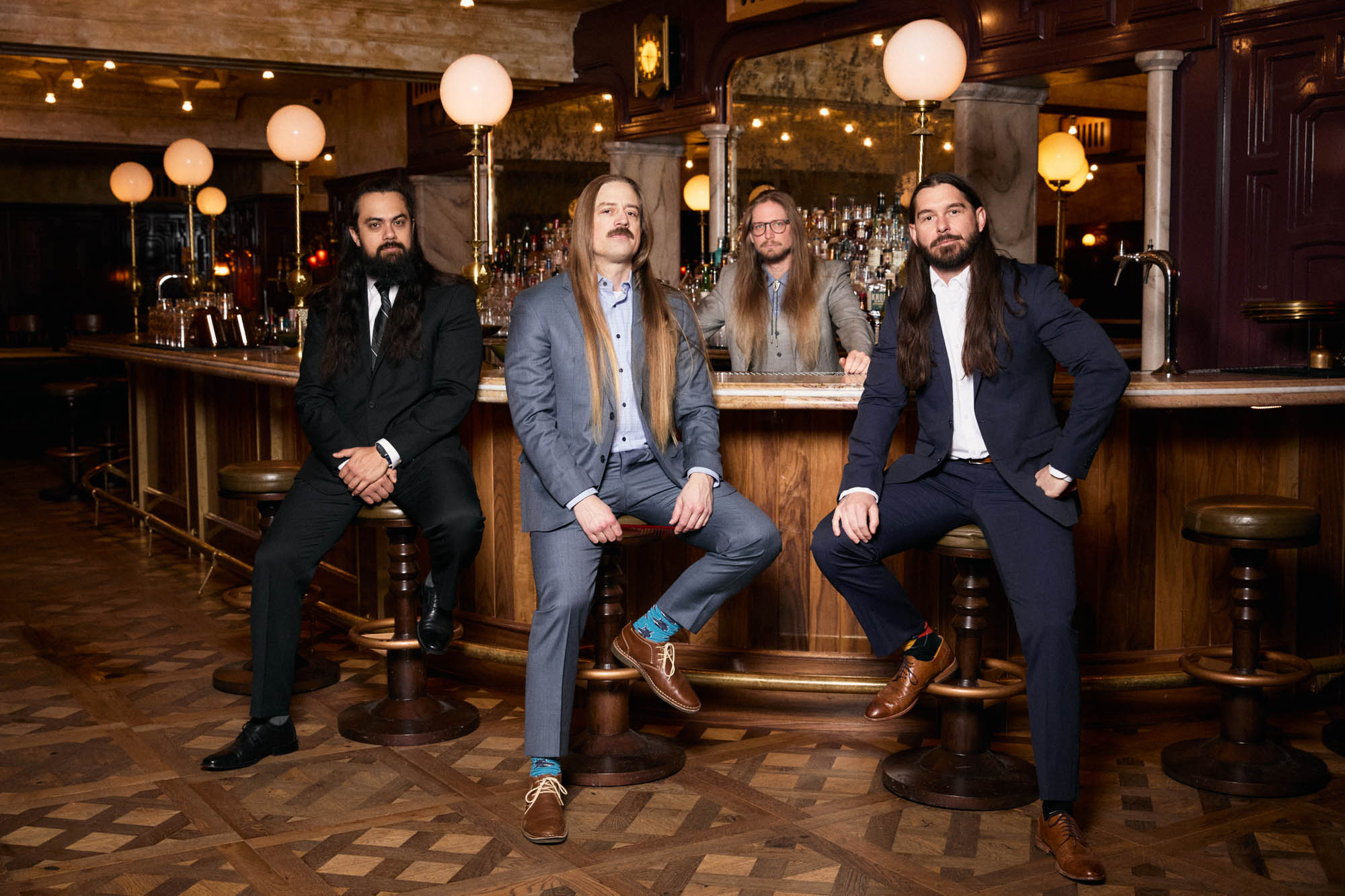 Jose Mejia, Chris Healy, Sean Horton, Lindsay Barto of The Longhairs, each wearing suits, sitting at an opulent bar, looking at the camera. By Erica Joan 2024.