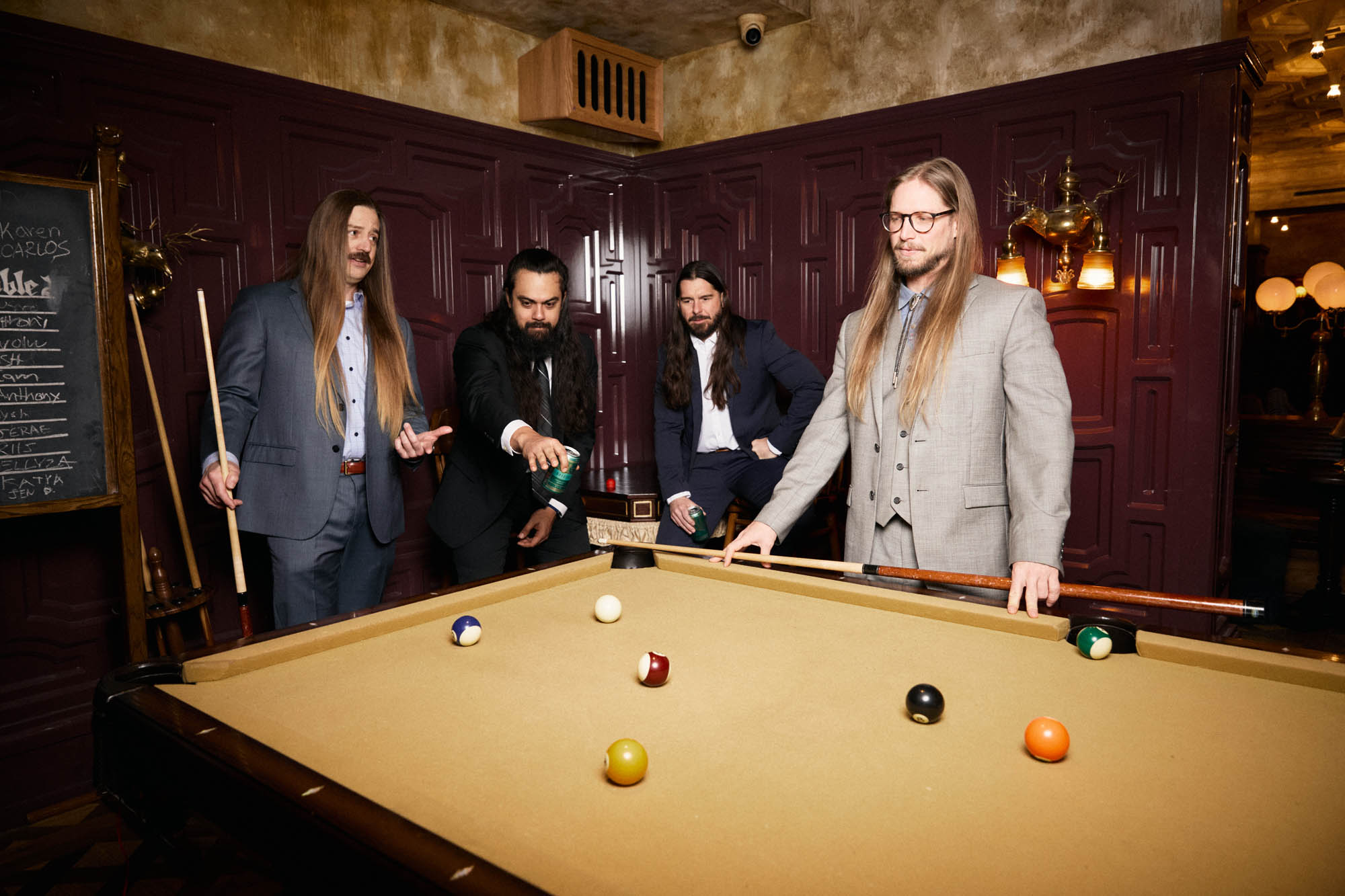 Chris Healy, Jose Mejia, Sean Horton, Lindsay Barto of The Longhairs, each wearing suits, standing around a pool table, looking down at the table. By Erica Joan 2024.