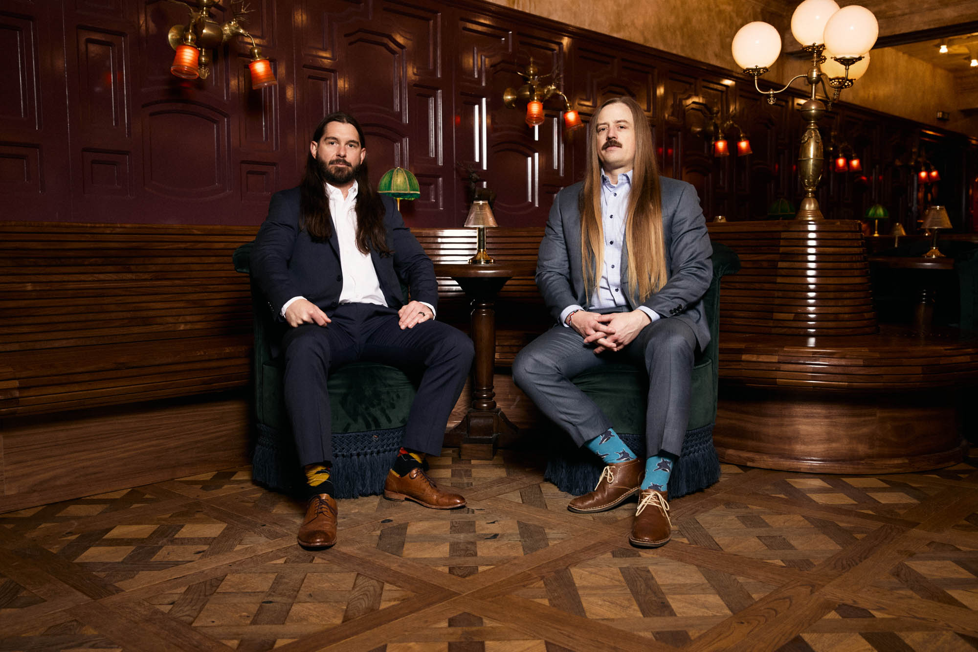 Lindsay Barto and Chris Healy, co-founders of The Longhairs, each wearing suits, sitting at a bar table in a fancy bar, looking at the camera. By Erica Joan 2024.
