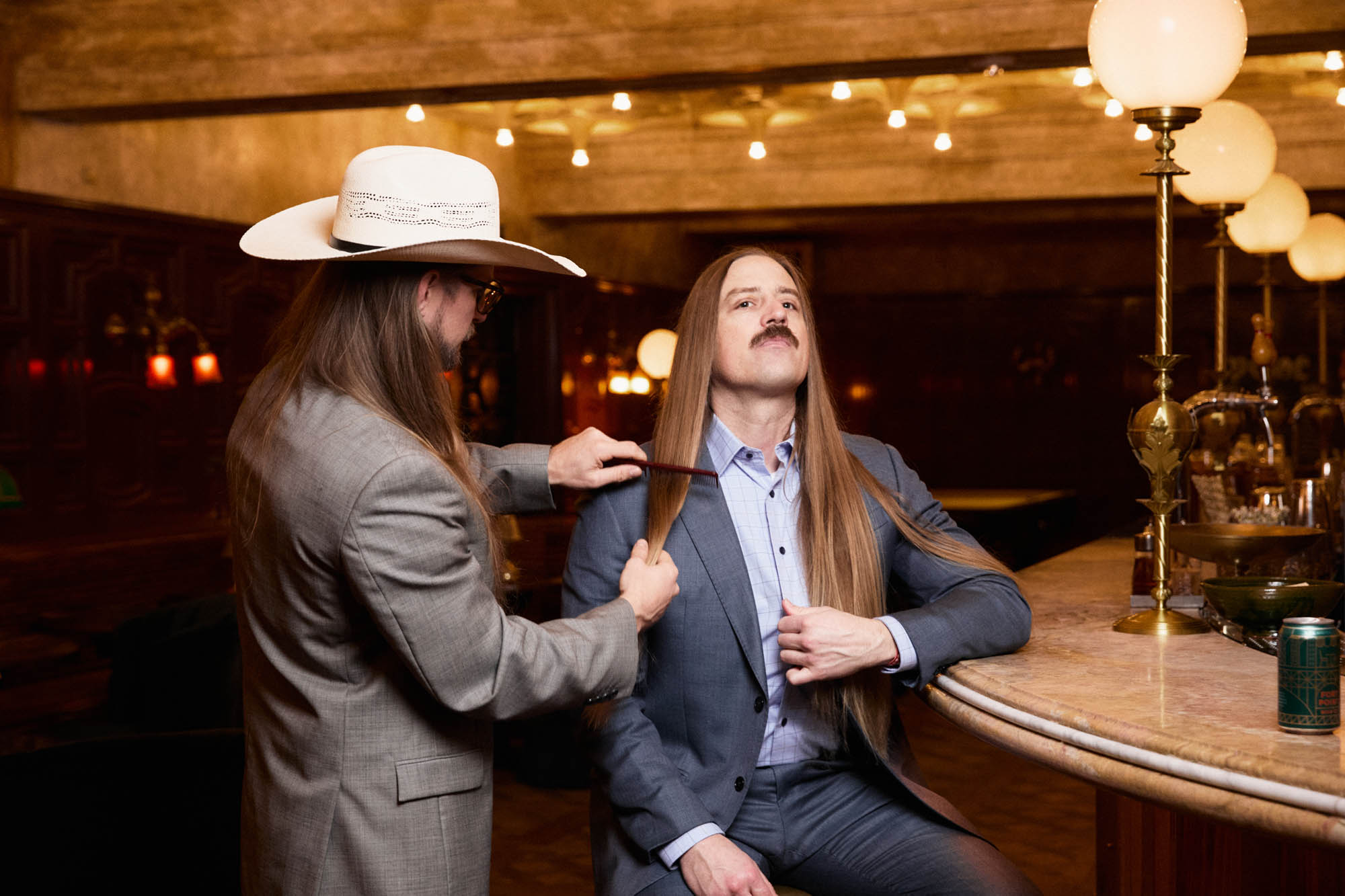 Sean Horton wearing a cowboy hat, passing a comb through Chris Healy’s long, blonde hair, each wearing suits, at an opulent bar, preparing for a picture. By Erica Joan 2024.