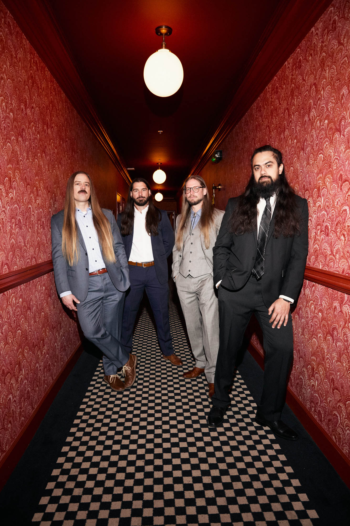 Chris Healy, Lindsay Barto, Sean Horton and Jose Mejia of The Longhairs, each wearing suits, standing in a hallway, looking casually at the camera. By Erica Joan 2024.