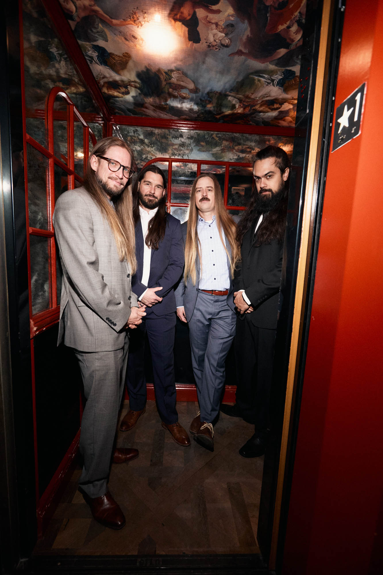 Sean Horton, Lindsay Barto, Chris Healy and Jose Mejia of The Longhairs, each wearing suits, standing in an elevator, looking at the camera, very cool. By Erica Joan 2024.
