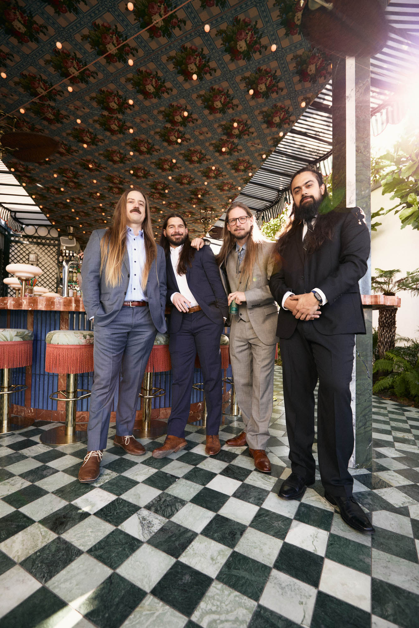 Chris Healy, Lindsay Barto, Sean Horton and Jose Mejia of The Longhairs, each wearing suits, standing at an outdoor bar, looking at the camera. By Erica Joan 2024.