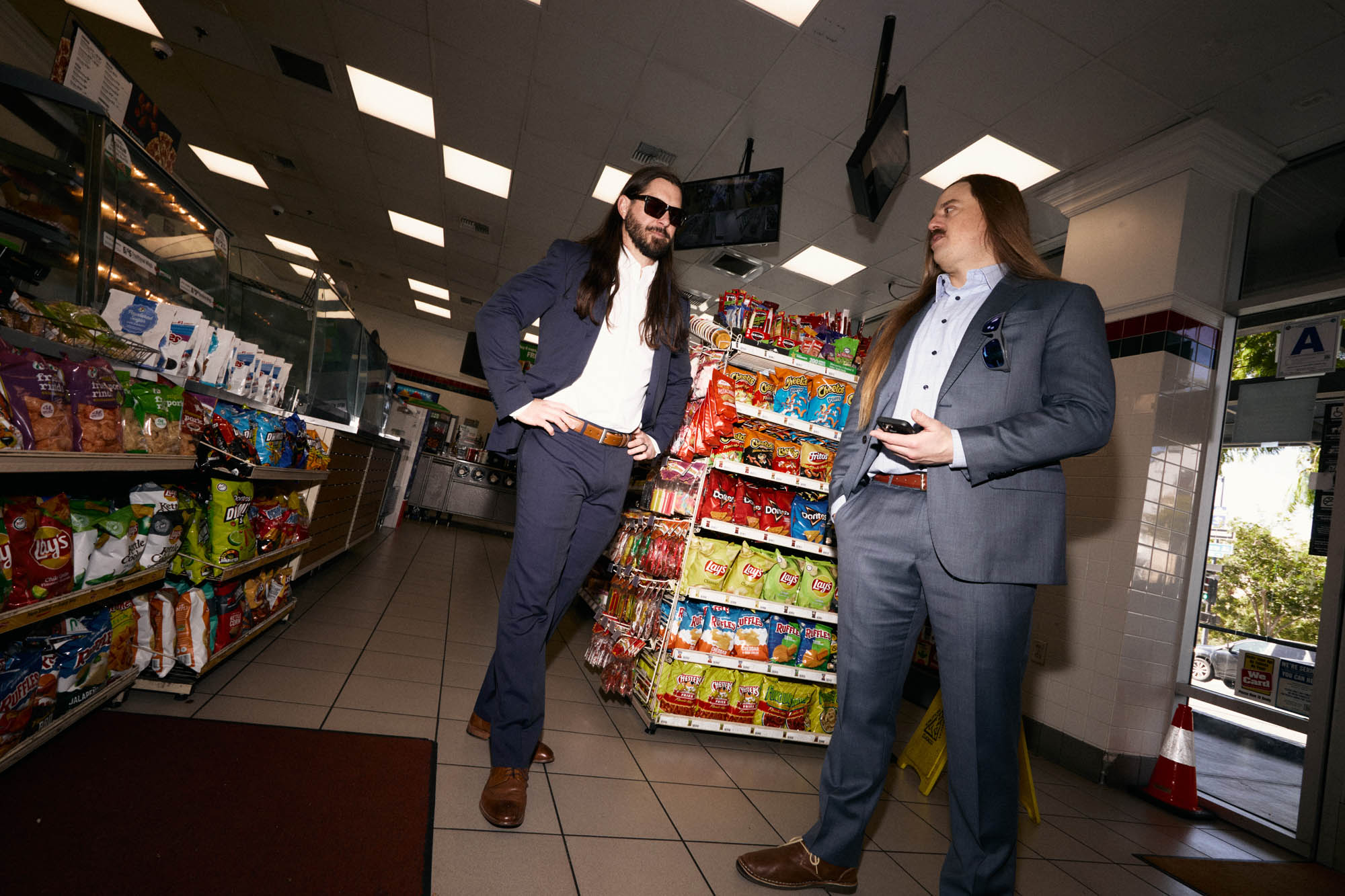 Lindsay Barto and Chris Healy of The Longhairs, each wearing suits, standing in a convenience store, deciding where to go next. By Erica Joan 2024.