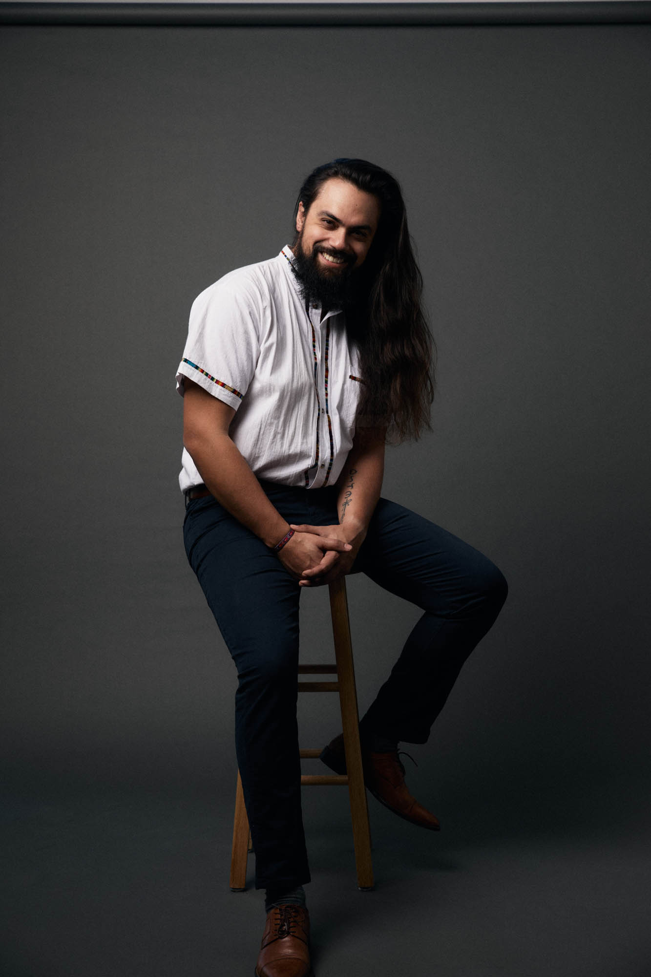 Jose Mejia smiling, sitting on a stool, and he’s a handsome devil. By Erica Joan 2024.