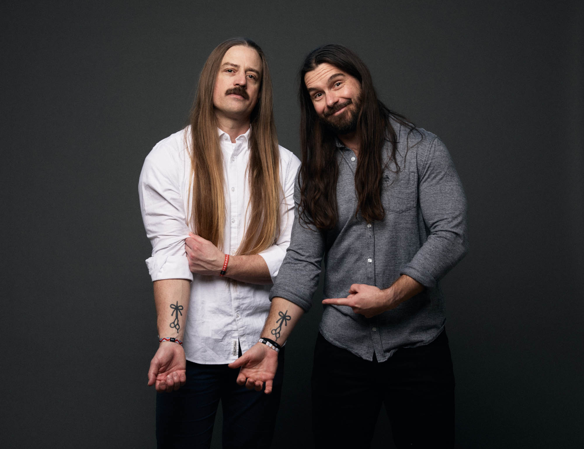 Chris Healy and Lindsay Barto, co-founders of The Longhairs, wearing button-up shirts, holding up their sleeves pointing at their tattoos of The Longhairs’ logo, looking at the camera. By Erica Joan 2024.