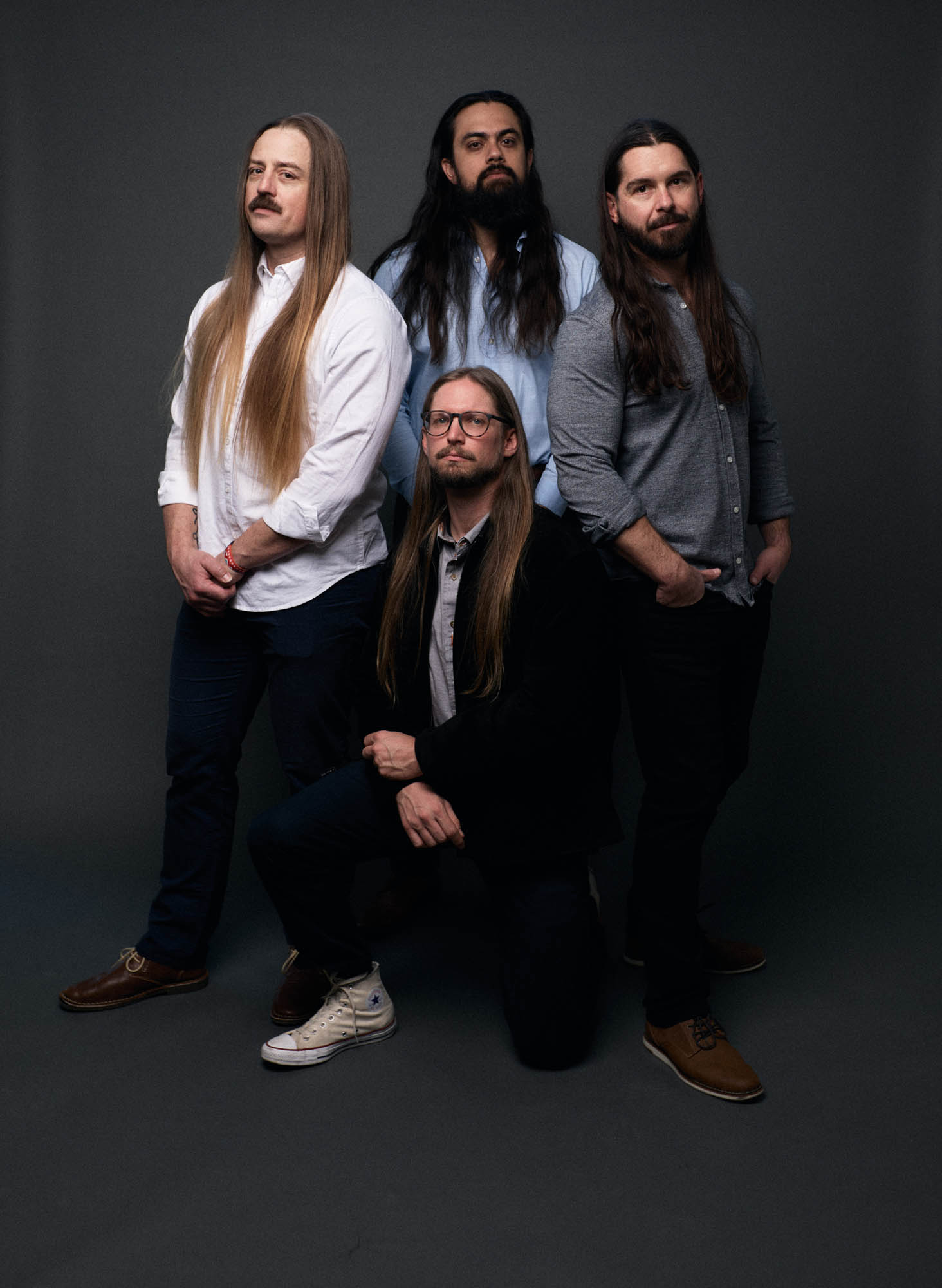 Chris Healy, Jose Mejia,  Lindsay Barto and Sean Horton (kneeling) of The Longhairs, in casual wear, in a studio, standing in diamond formation looking at the camera with straight faces. By Erica Joan 2024.